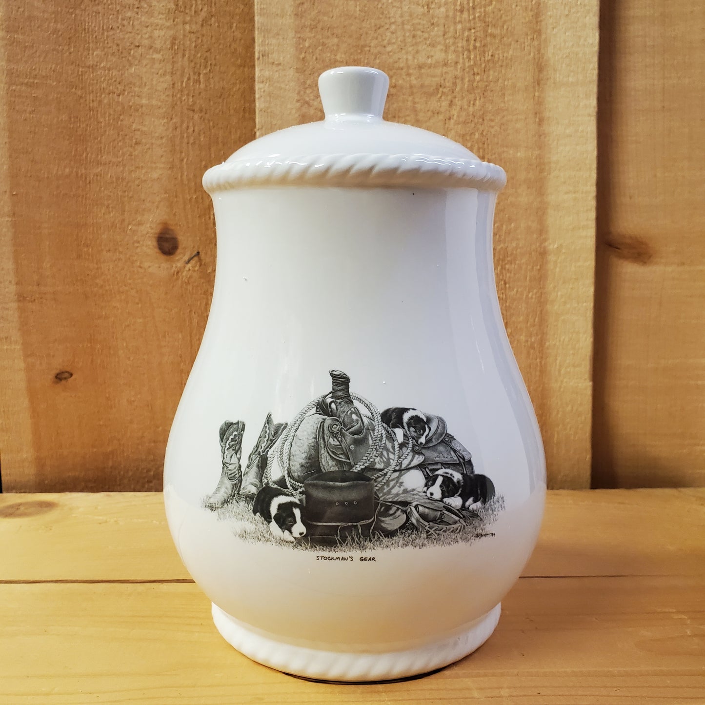 A white cookie jar with a lid, featuring a black and gray pencil art depiction of puppies on a western saddle