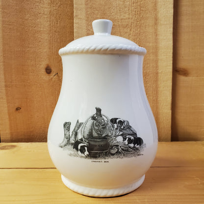 A white cookie jar with a lid, featuring a black and gray pencil art depiction of puppies on a western saddle