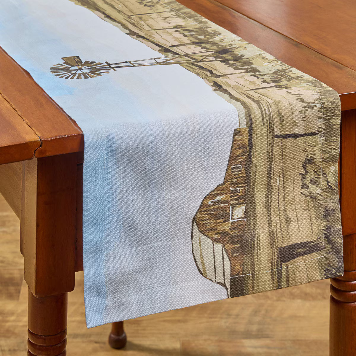 a table runner with a prairie scene containing a windmill and an old barn on a wooden table