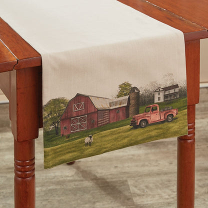 Table runner with a farm scene featuring a red barn and rusty truck on a wooden table.