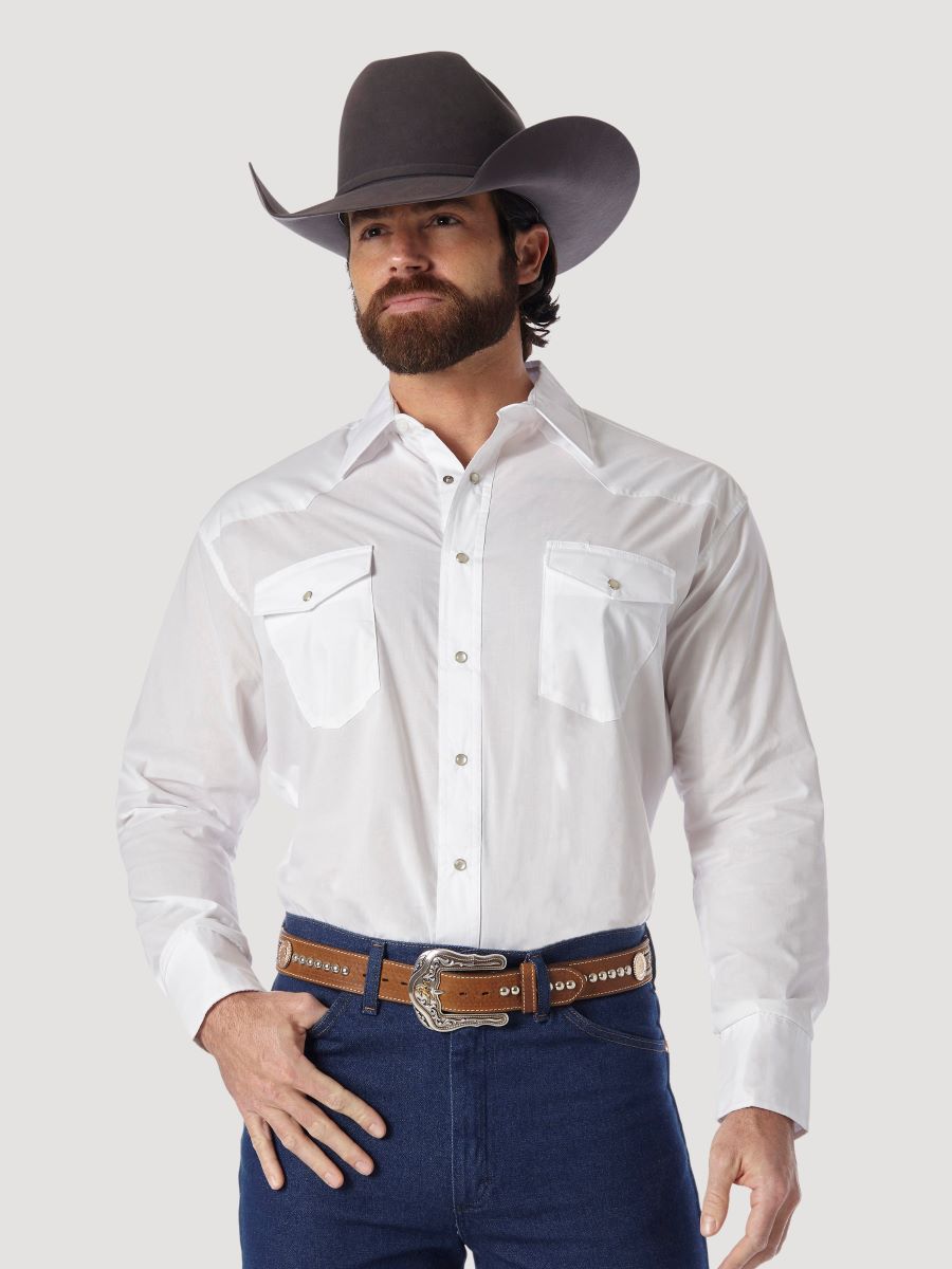 A man wearing a white long-sleeve Wrangler western shirt with two chest pockets, a brown belt, and blue jeans, standing against a light gray background.