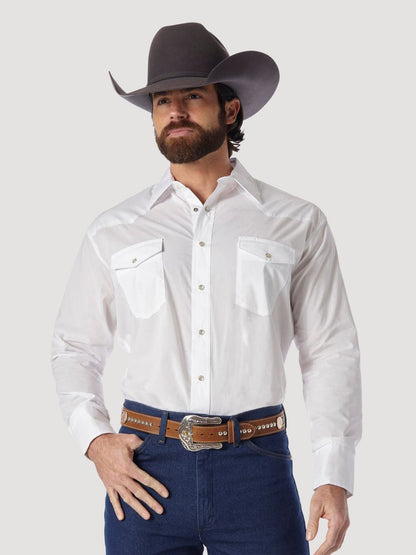 A man wearing a white long-sleeve Wrangler western shirt with two chest pockets, a brown belt, and blue jeans, standing against a light gray background.