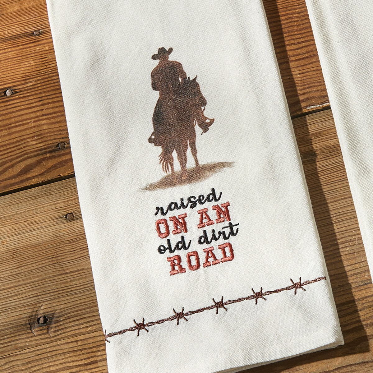 Towel with cowboy graphic and 'raised on an old dirt road' text on a wooden surface