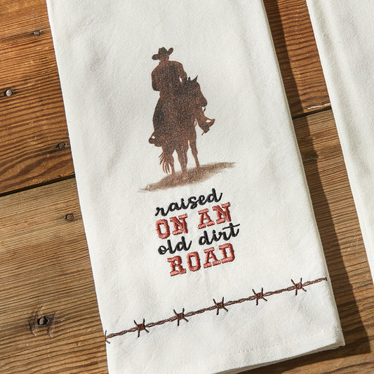 Towel with cowboy graphic and 'raised on an old dirt road' text on a wooden surface
