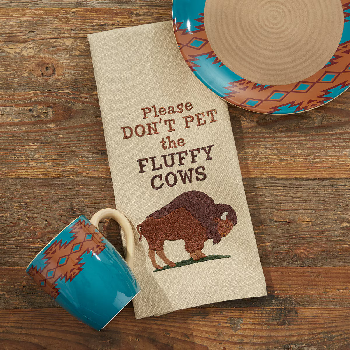 a kitchen towel that has a bison and the words 'please don't pet the fluffy cows' on a wooden background with a plate and coffee cup around it