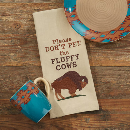 a kitchen towel that has a bison and the words 'please don't pet the fluffy cows' on a wooden background with a plate and coffee cup around it