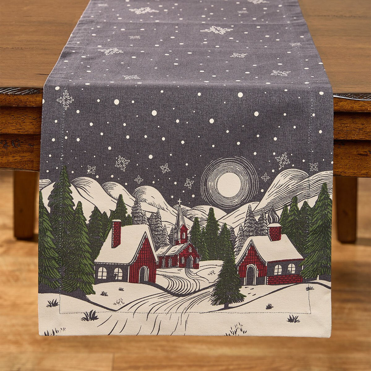 A fabric table runner from Park Designs laid across a wooden table, printed with a winter village scene featuring snow-covered roofs, evergreen trees, mountains, a moon, and a starry night sky.