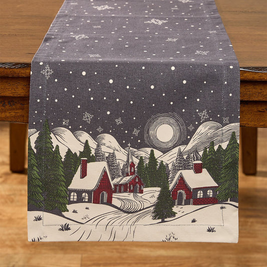 A fabric table runner from Park Designs laid across a wooden table, printed with a winter village scene featuring snow-covered roofs, evergreen trees, mountains, a moon, and a starry night sky.