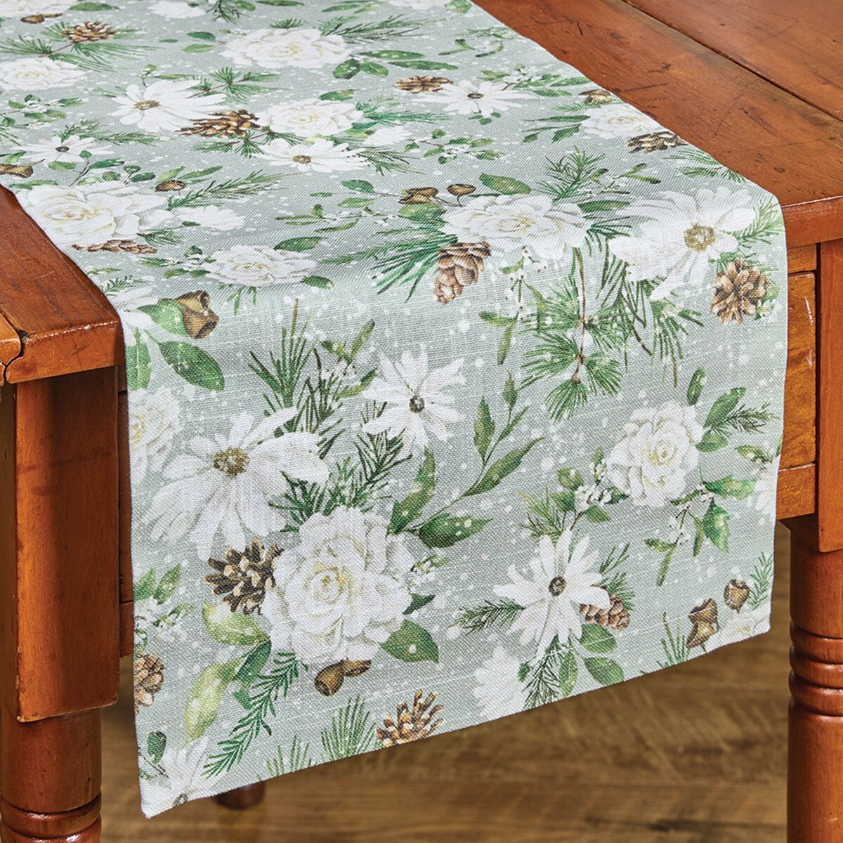 A floral table runner from Park Designs draped over a wooden table, featuring white flowers, green pine branches, and brown pinecones on a light gray background.
