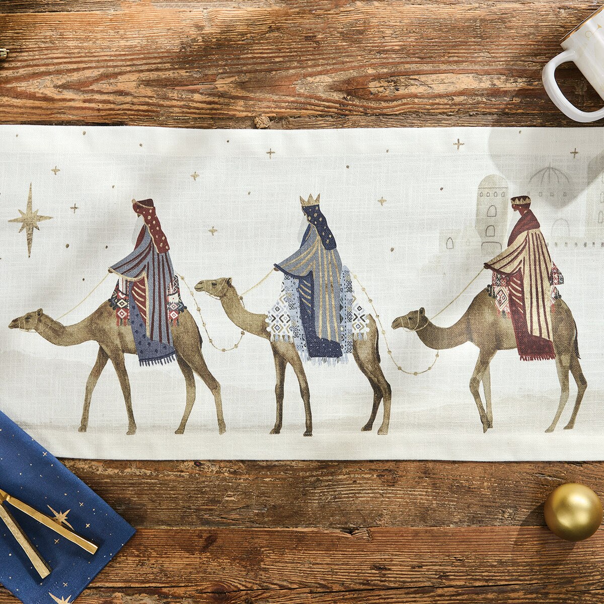 A rectangular table runner from Park Designs with a Christmas Nativity scene print, laid flat on a rustic wooden table. The runner features three camels carrying figures in robes under a star motif.