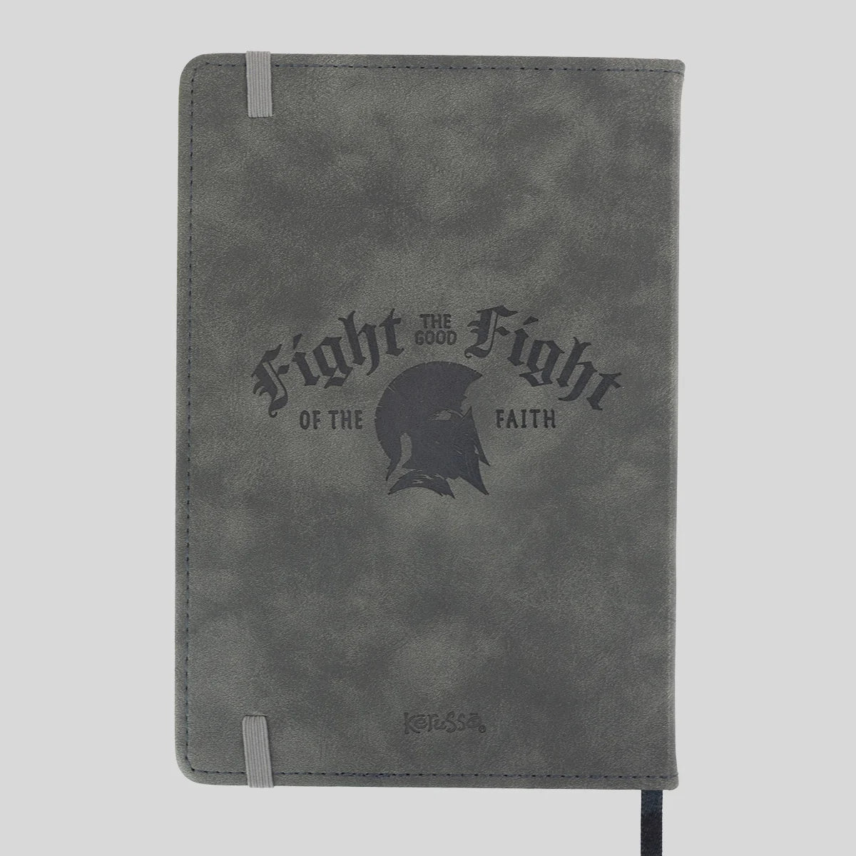 A gray textured Kerusso notebook or journal with a dark gray printed graphic on the front that reads 'Fight for the Good Fight of the Faith' around a helmet silhouette. It has a gray elastic closure band and a black ribbon bookmark hanging from the bottom right, shown against a light gray background.