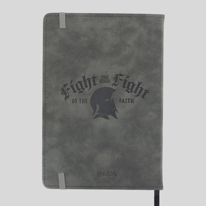 A gray textured Kerusso notebook or journal with a dark gray printed graphic on the front that reads 'Fight for the Good Fight of the Faith' around a helmet silhouette. It has a gray elastic closure band and a black ribbon bookmark hanging from the bottom right, shown against a light gray background.