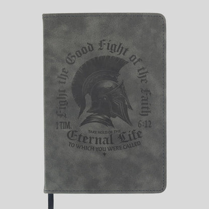 A gray faux-leather journal from Kerusso featuring a black embossed design of a Spartan helmet and the text 'Fight the Good Fight of the Faith' and 'Eternal Life' from 1 Timothy 6:12.