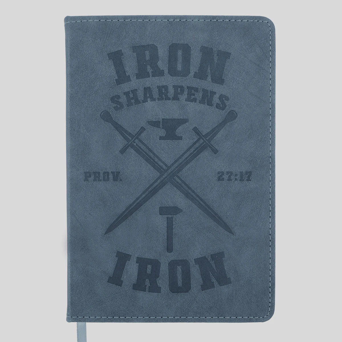 A blue-gray faux-leather journal from Kerusso with a dark gray embossed design featuring the words 'IRON SHARPENS', crossed swords, an anvil, and 'IRON' with a hammer. A white ribbon bookmark is visible at the bottom left.