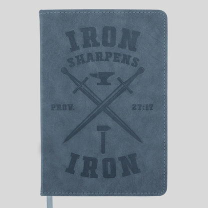 A blue-gray faux-leather journal from Kerusso with a dark gray embossed design featuring the words 'IRON SHARPENS', crossed swords, an anvil, and 'IRON' with a hammer. A white ribbon bookmark is visible at the bottom left.