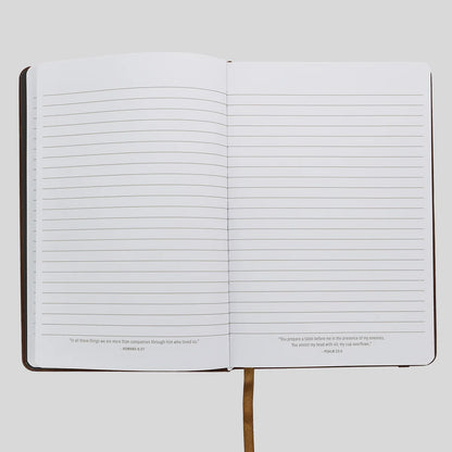 An open lined journal or notebook from Kerusso with a dark cover, blank horizontal ruled pages, and small Bible verse quotes printed at the bottom of each page, shown on a light gray background.