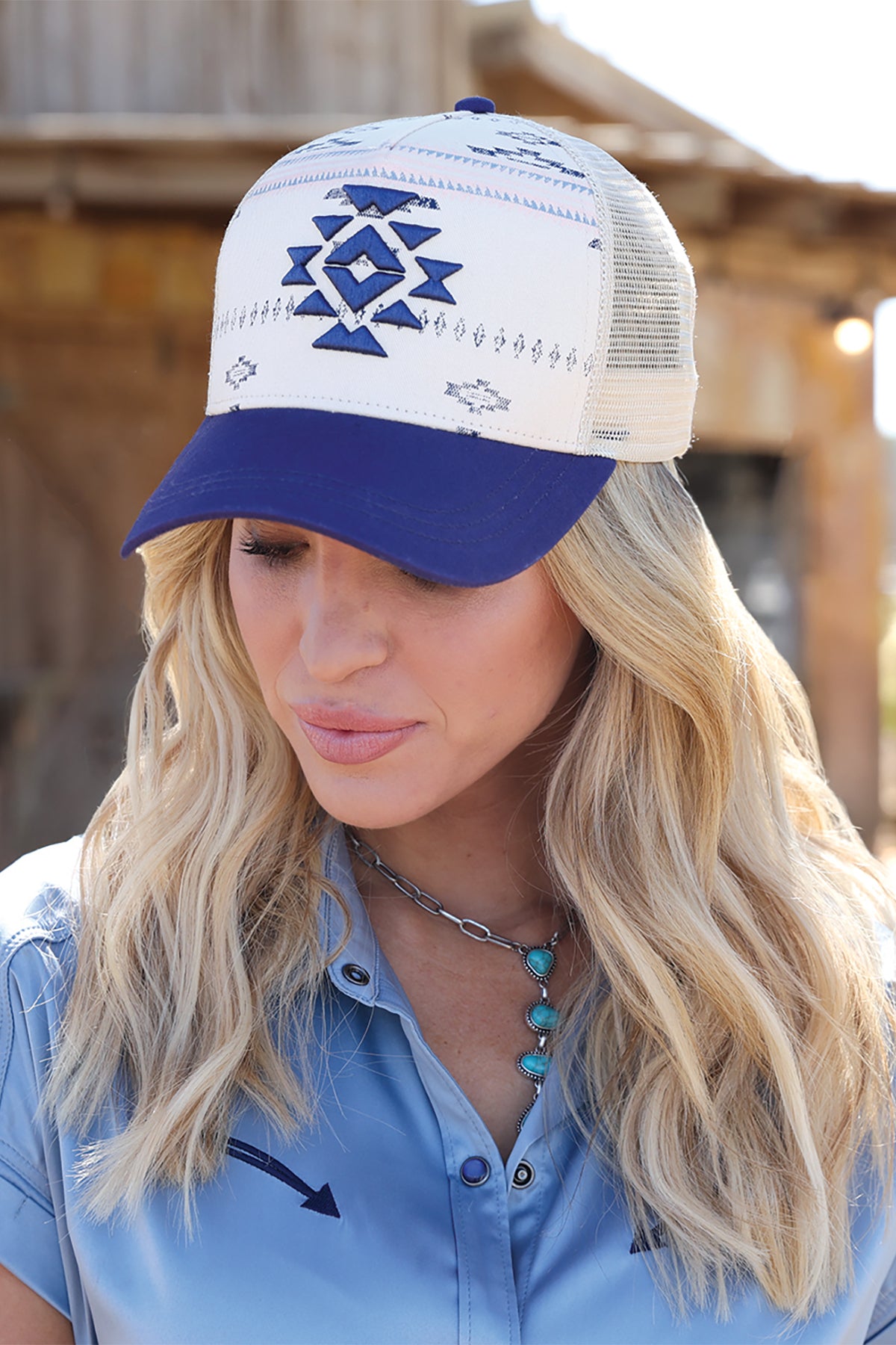 Woman wearing a white and blue cruel denim cap with a geometric design, standing in front of a wooden structure.