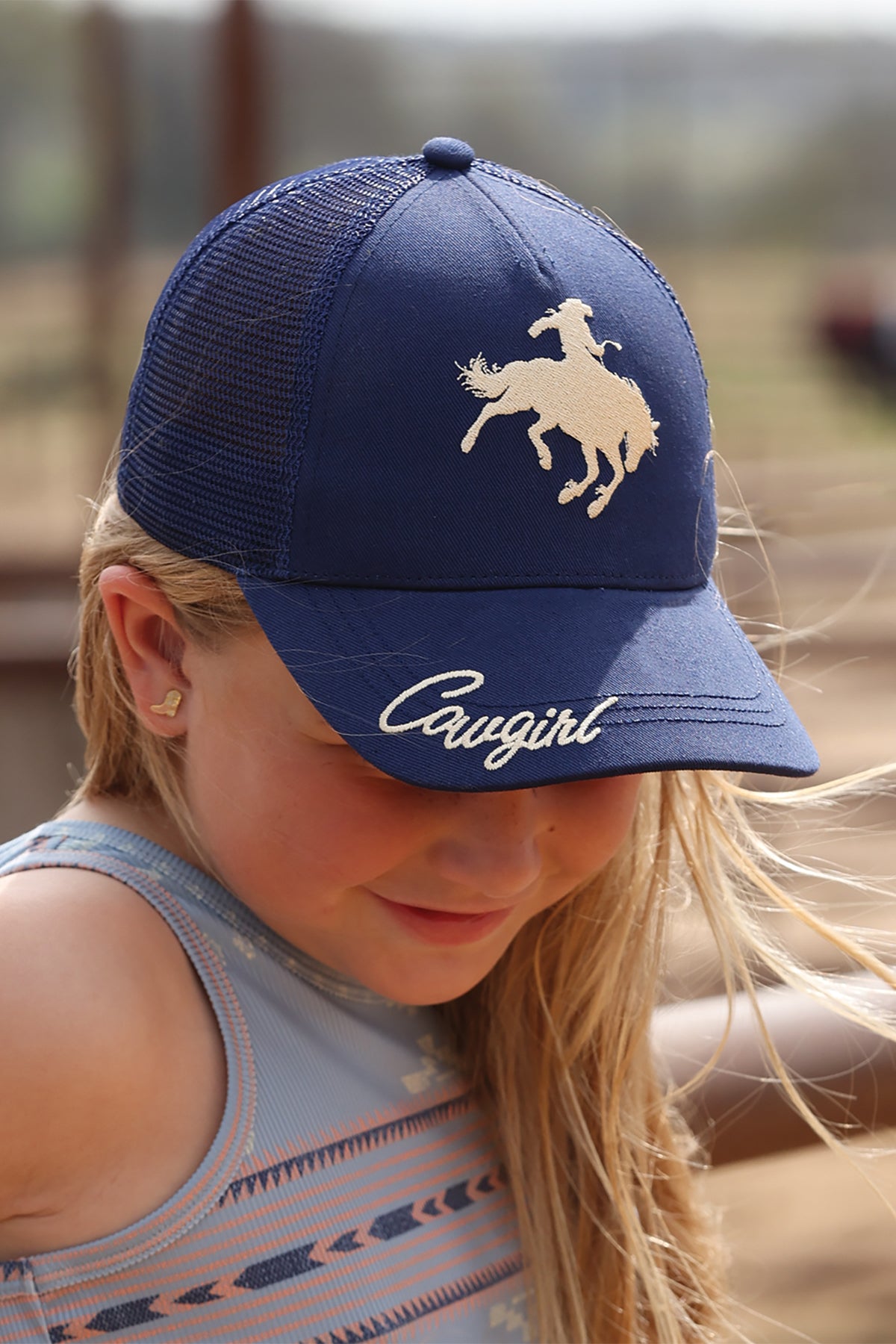 Child wearing a blue cap with a horse logo and 'Cowgirl' text, outdoors.