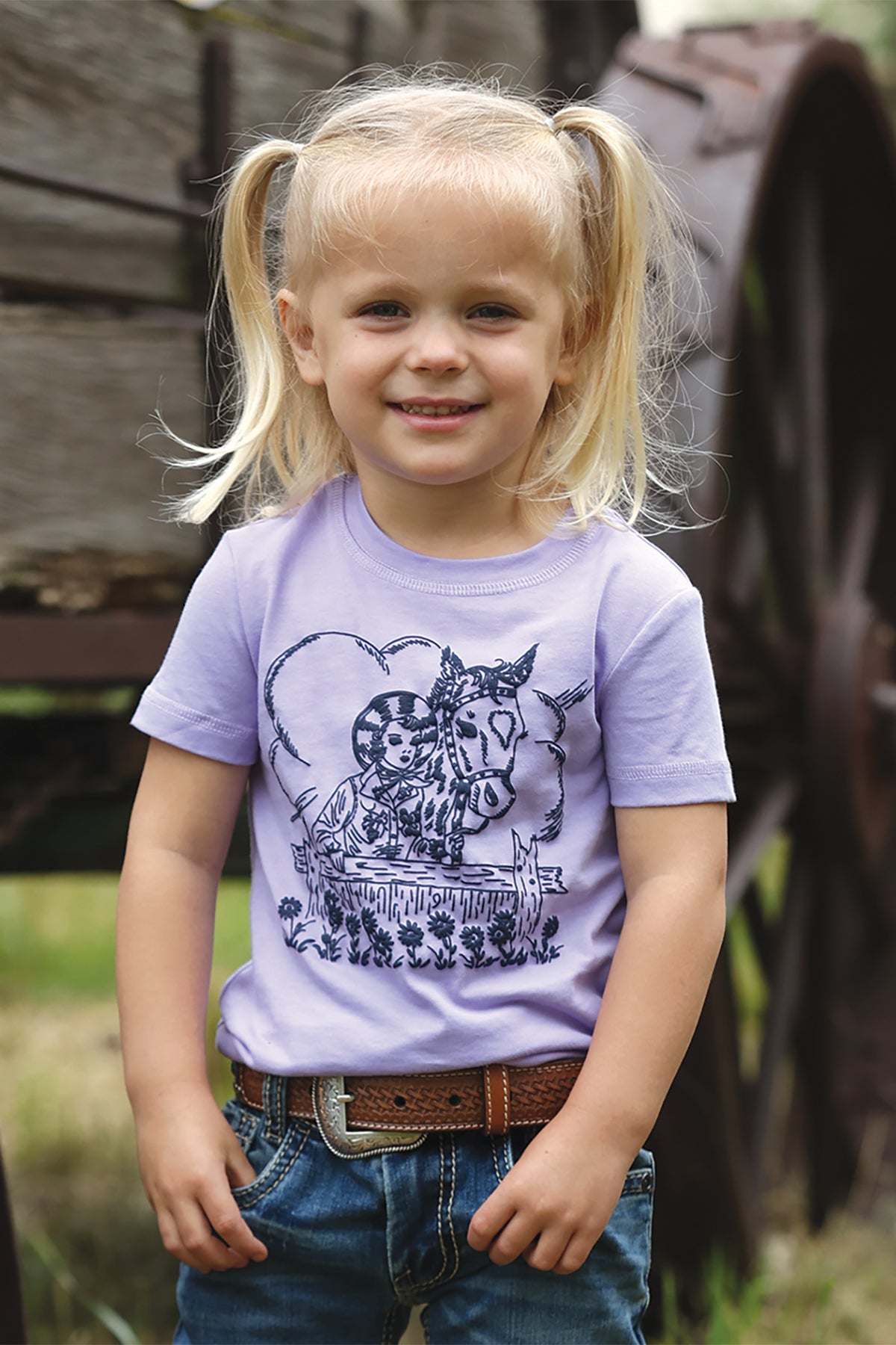 Young girl wearing a purple Cinch  t-shirt with a horse graphic, standing in front of a wooden structure.