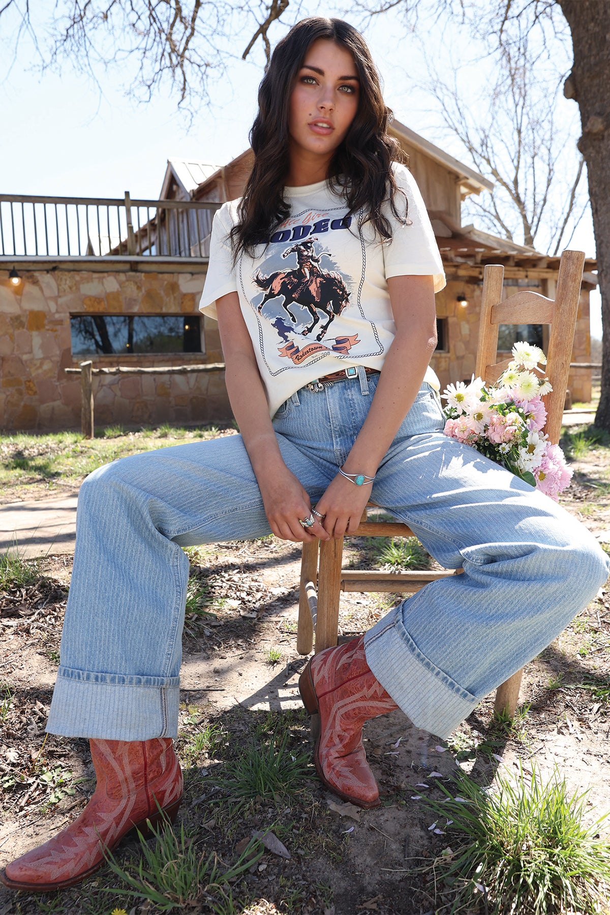 Woman sitting outdoors wearing a graphic t-shirt, jeans, and cowboy boots.