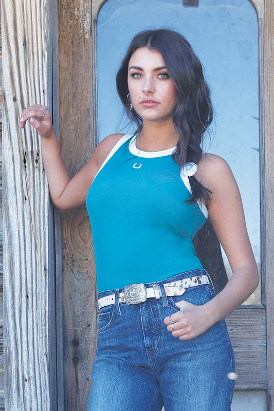 Woman wearing a teal tank top by Cruel Denim and jeans standing against a wooden wall.