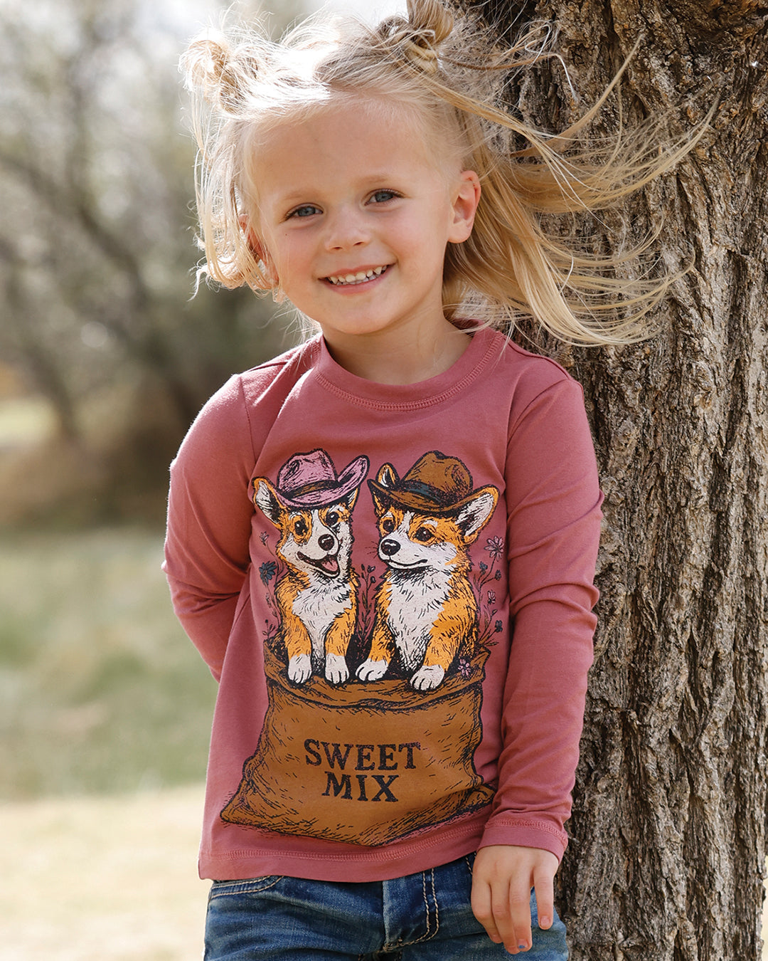 Girl wearing a pink Cruel Girls shirt with some puppies in a feed bag wearing cowboy hats, leaning against a tree