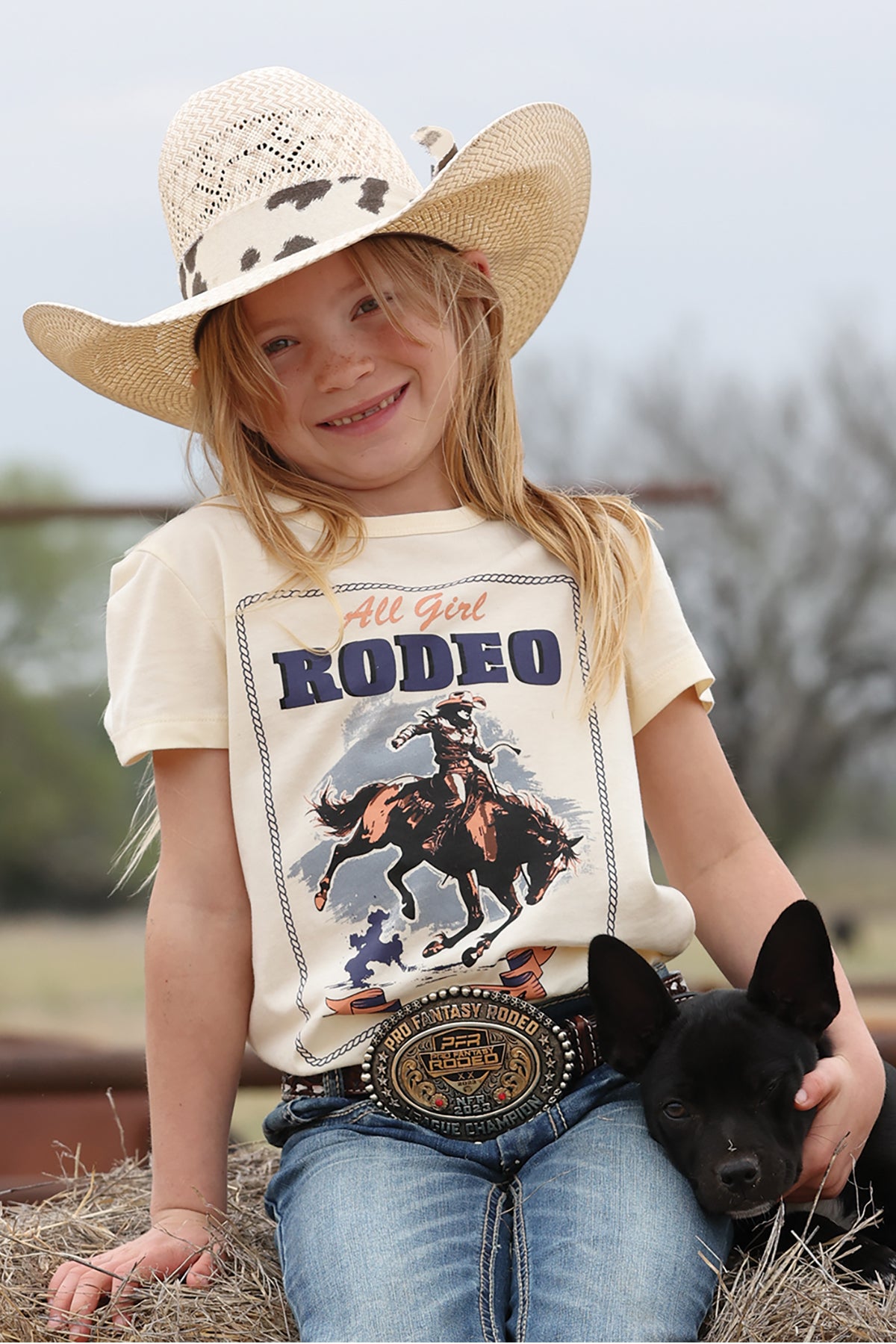 Young girl wearing a 'All Girl Rodeo' cruel girl t-shirt with a cowboy graphic, holding a black dog, and wearing a cowboy hat.