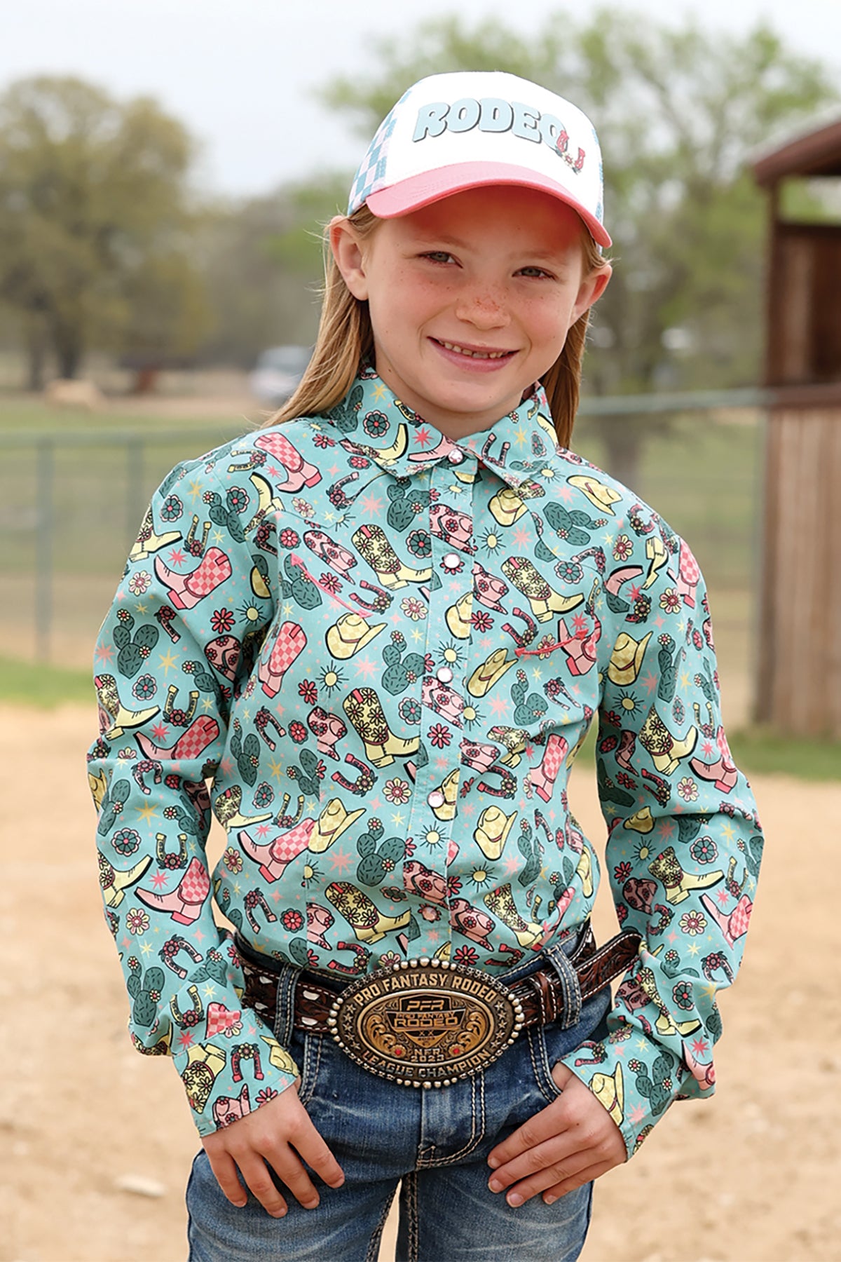 Young girl wearing a colorful patterned Cruel Girl shirt with a cap, standing outdoors.