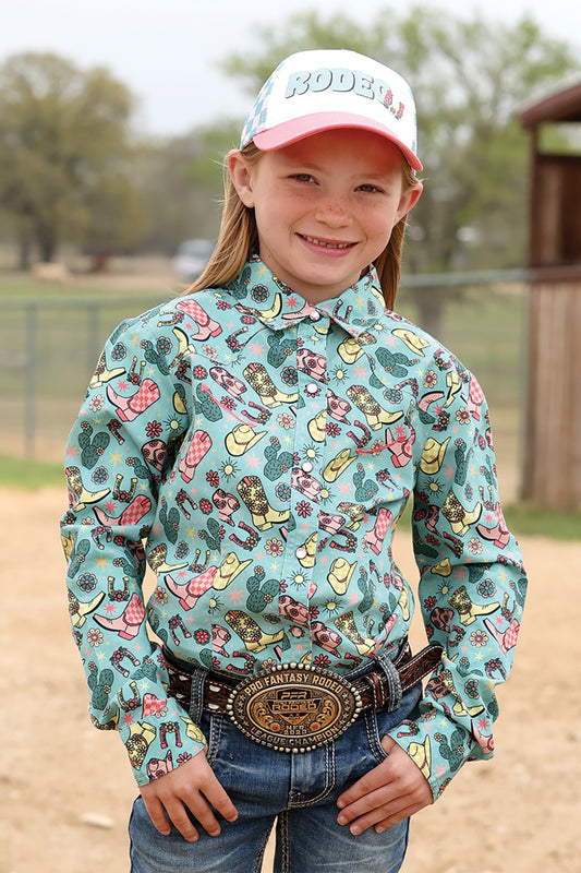 Young girl wearing a colorful patterned Cruel Girl shirt with a cap, standing outdoors.