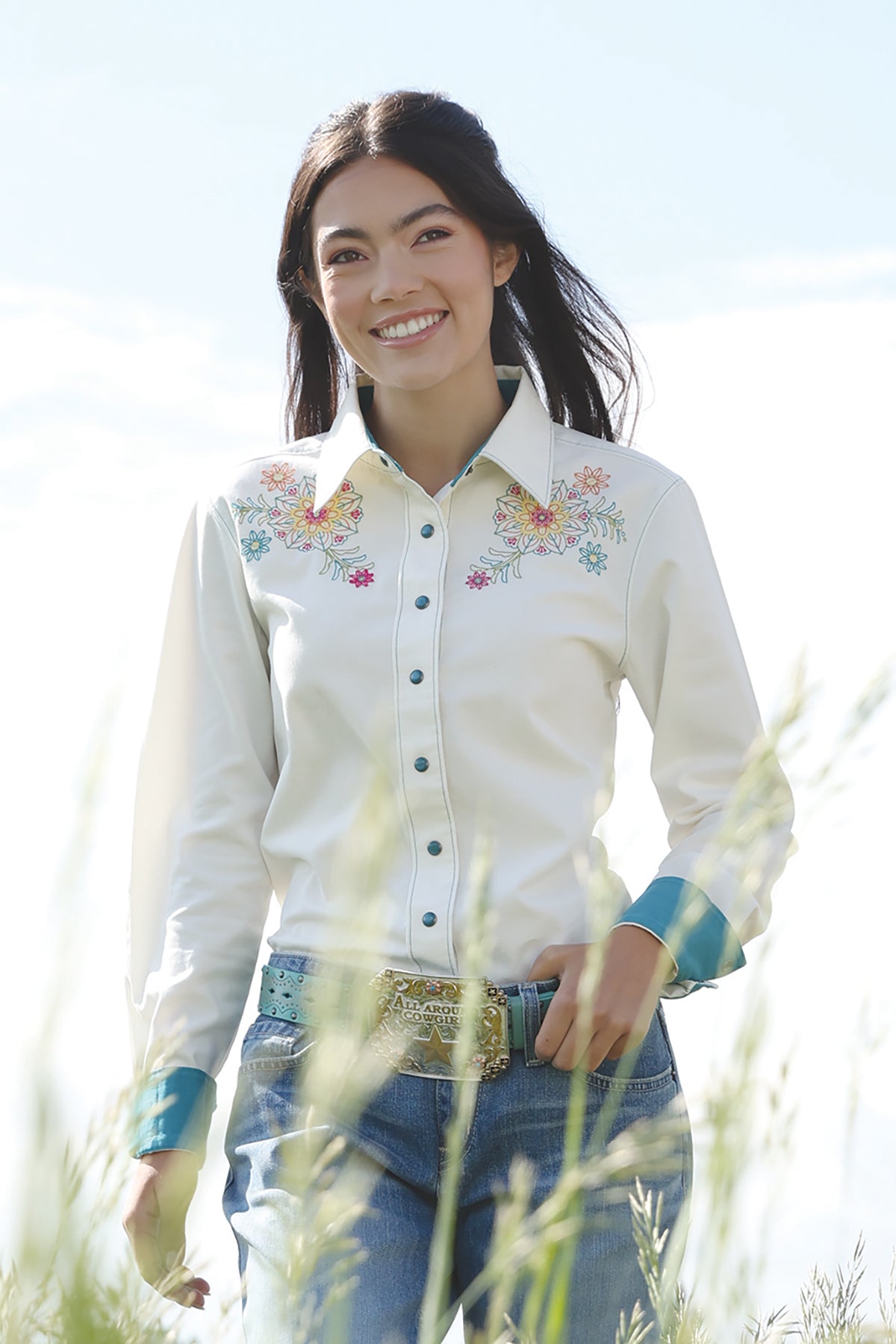 Woman wearing Cinch shirt with flowers on it and blue jeans and buckle in a field