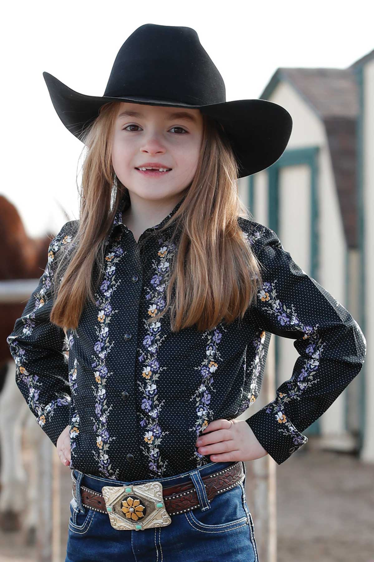 Girl wearing a black floral Cruel Girls shirt, blue jeans and a black cowboy hat standing in a farm yard