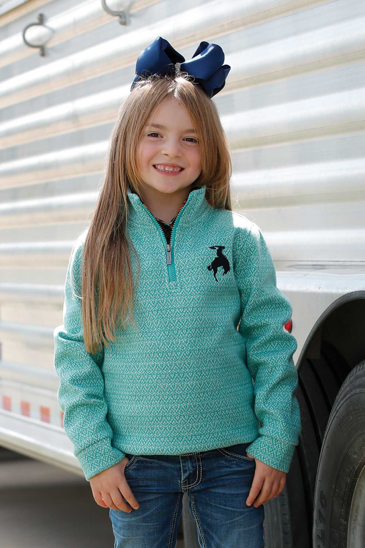 Young girl wearing a teal sweater with a logo, standing in front of a trailer.