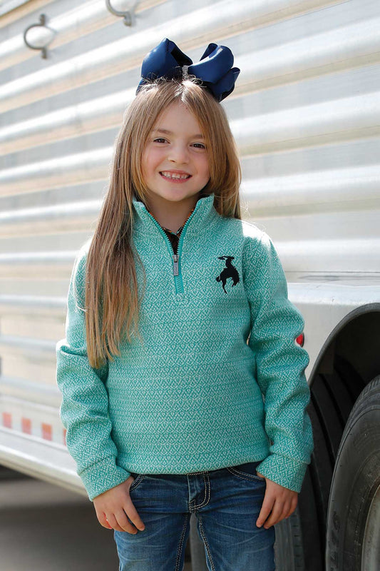 Young girl wearing a teal sweater with a logo, standing in front of a trailer.