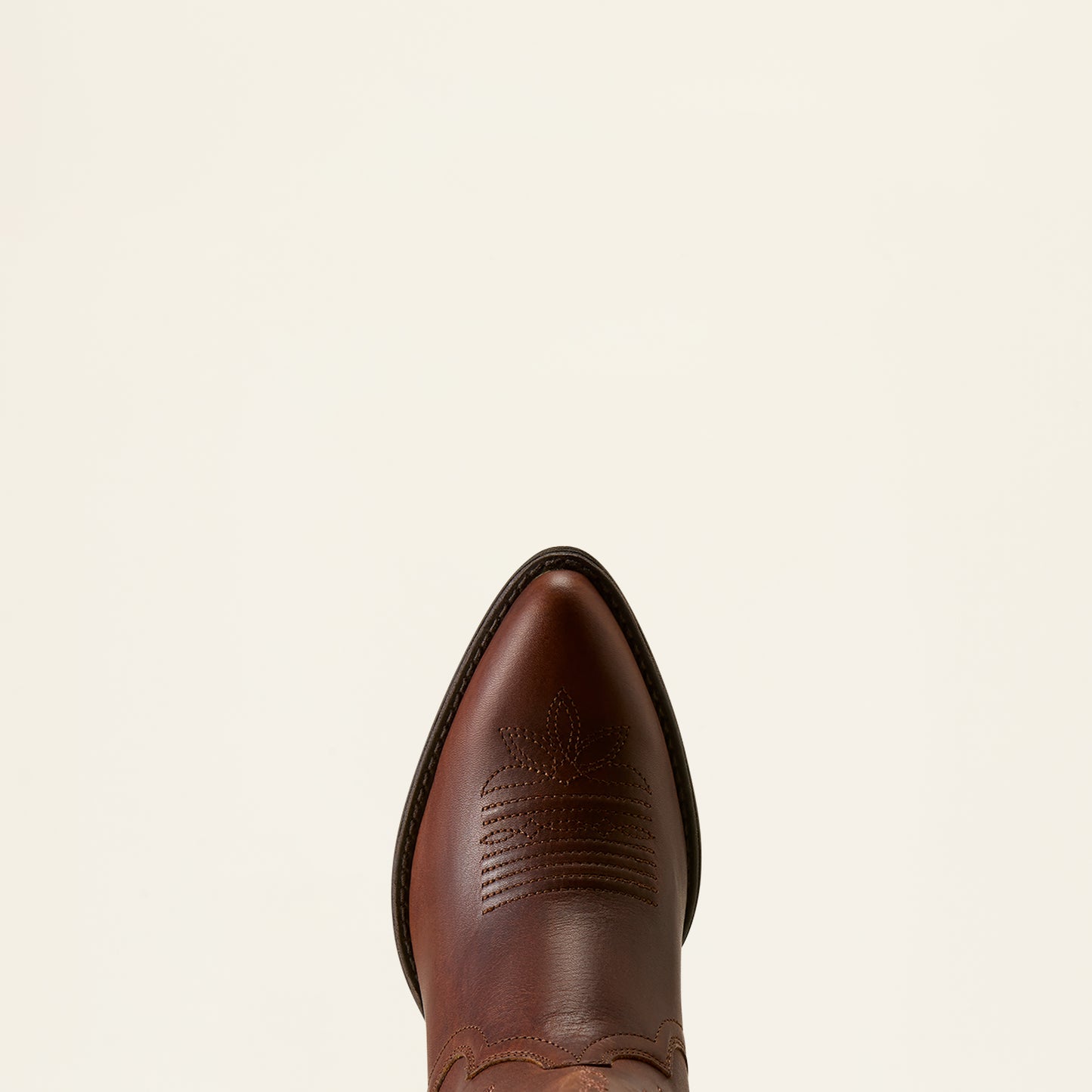 A close-up top-down view of a brown leather cowboy boot toe, showing the pointed tip and decorative stitching, set against a solid off-white background.