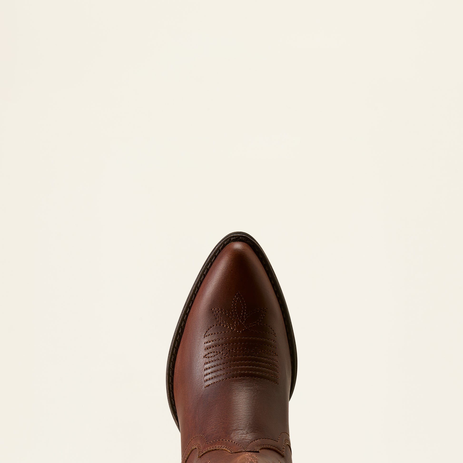 A close-up top-down view of a brown leather cowboy boot toe, showing the pointed tip and decorative stitching, set against a solid off-white background.
