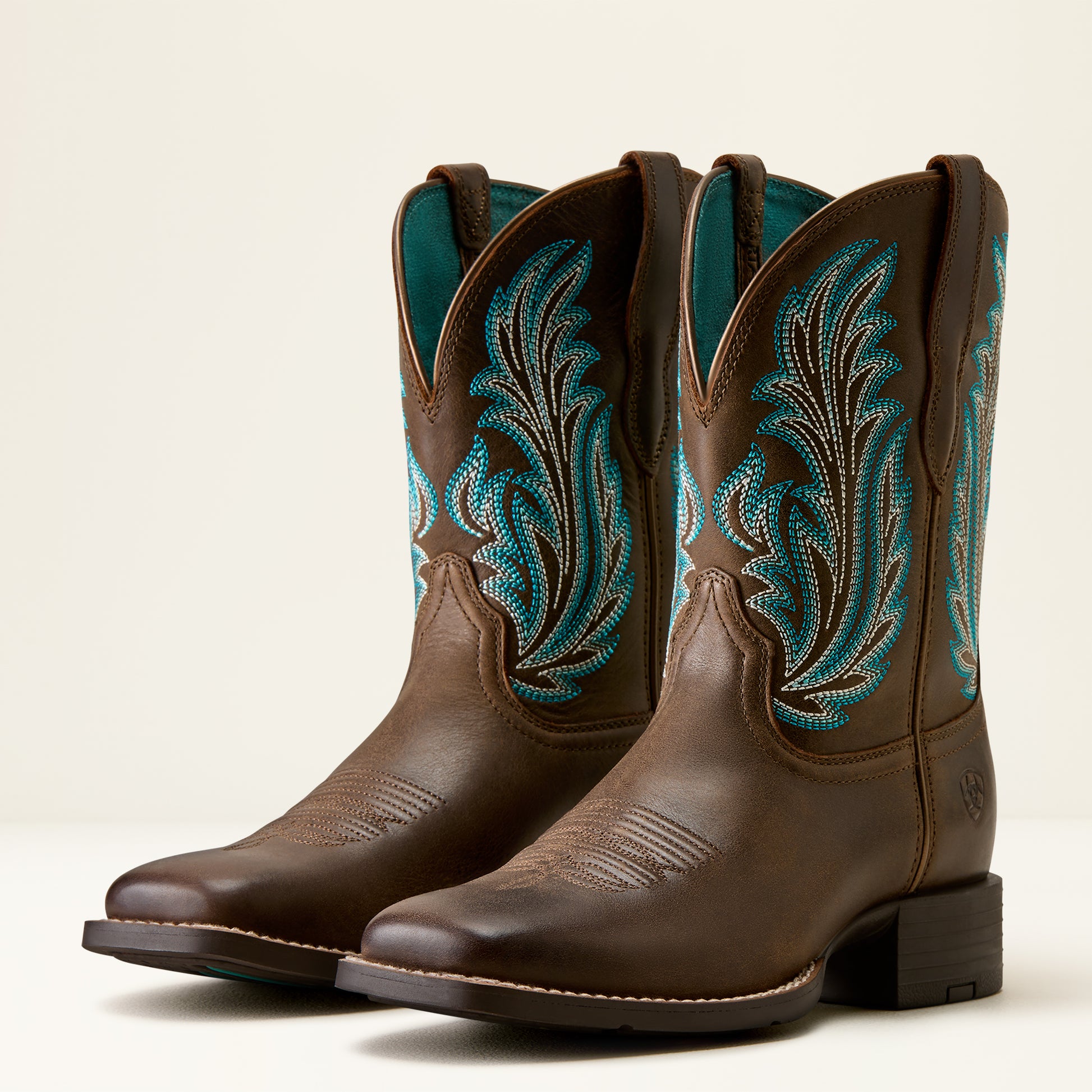A pair of brown leather cowboy boots with turquoise and white embroidered floral patterns on the shafts, shown against a plain light beige background.