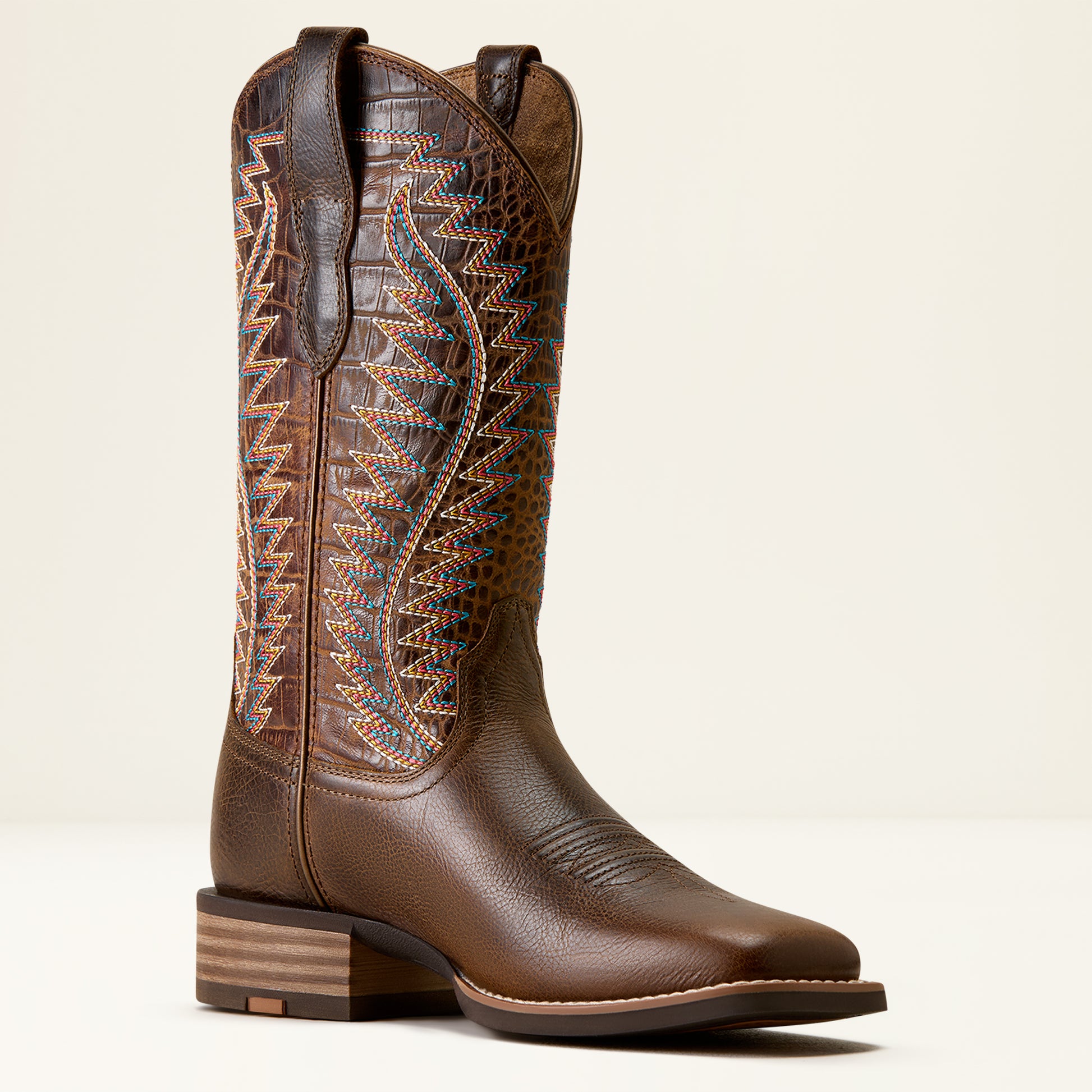 A single brown leather cowboy boot with a square toe, stacked wood-look heel, and intricate multicolor embroidered stitching on the shaft, shown against a plain light beige background.