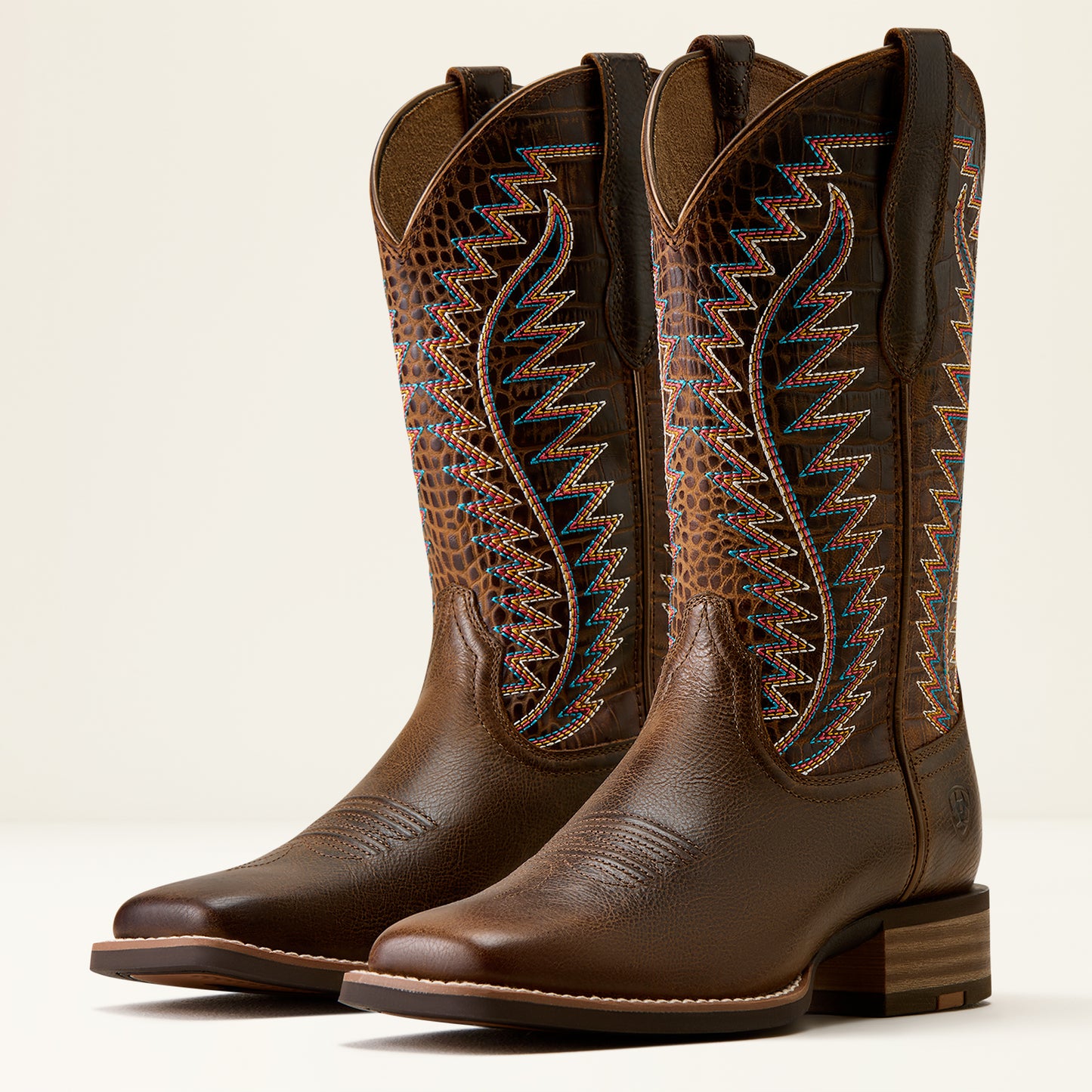 A pair of brown leather cowboy boots with decorative embroidered shafts in blue, orange, and gold, shown on a light beige background.