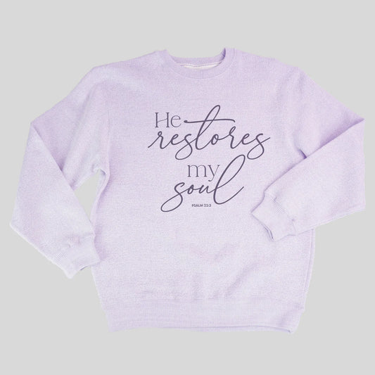 A light lavender crewneck sweatshirt from Grace & Truth featuring the printed text 'He restores my soul' in black script and serif fonts, with 'PSALM 23:3' printed below in a small block font, displayed on a solid light gray background.