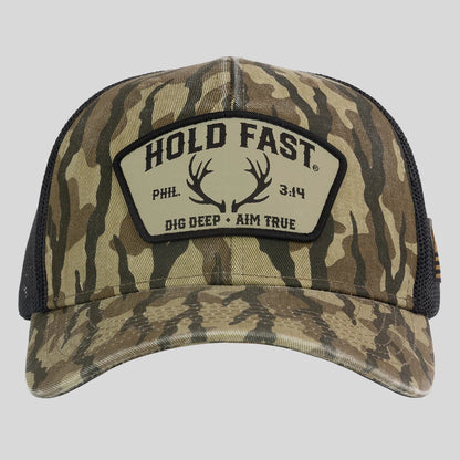 A camouflage trucker hat from Hold Fast with a black mesh back and a front patch that reads 'HOLD FAST' with antlers and 'DIG DEEP • AIM TRUE'.