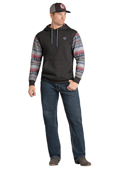 Man wearing a black hoodie with patterned sleeves and relaxed fit Hooey blue jeans on a white background