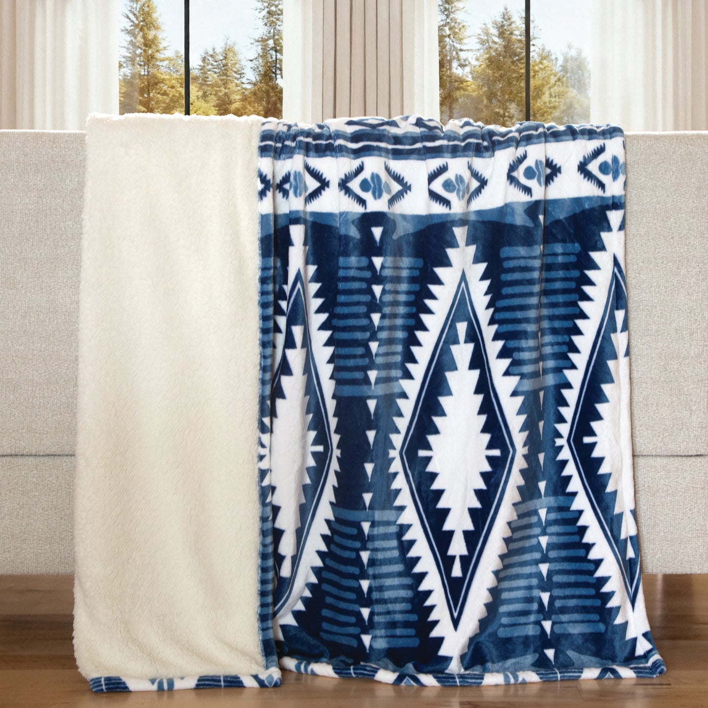 A plush throw blanket draped over a sofa, showing a cream sherpa-like reverse side and a blue-and-white Southwestern geometric patterned front. The sofa is in front of large windows with trees visible outside.
