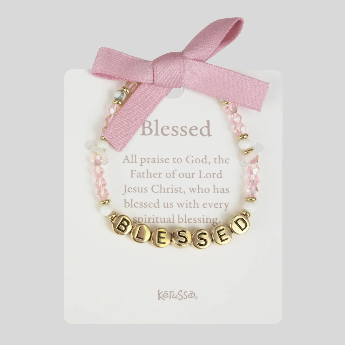 A pink and gold beaded bracelet with a 'BLESSED' lettered charm, presented on a white card with a pink ribbon bow. The card features the text 'Blessed' and the brand name 'Kerusso'.