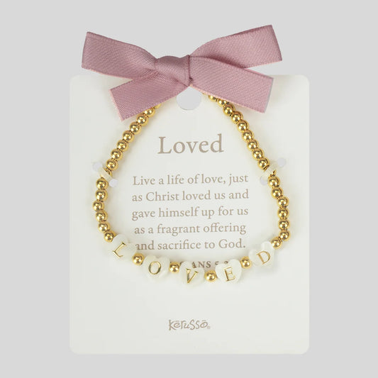 A gold beaded bracelet with white letter beads spelling 'LOVED' and small gold beads, displayed on a white card with a pink ribbon bow. The card reads 'Loved' with a Bible verse and the brand name 'Kerusso' at the bottom.