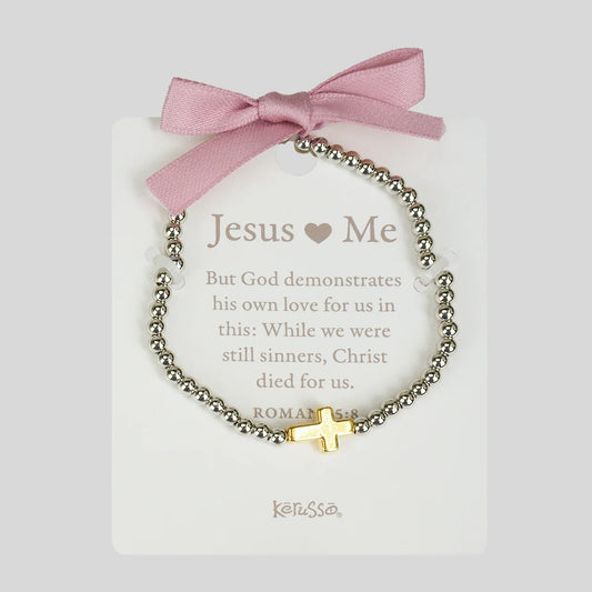 A beaded bracelet with a gold cross charm and white heart beads, displayed on a white card with a pink ribbon bow. The card reads 'Jesus ♥ Me' and shows the brand Kerusso