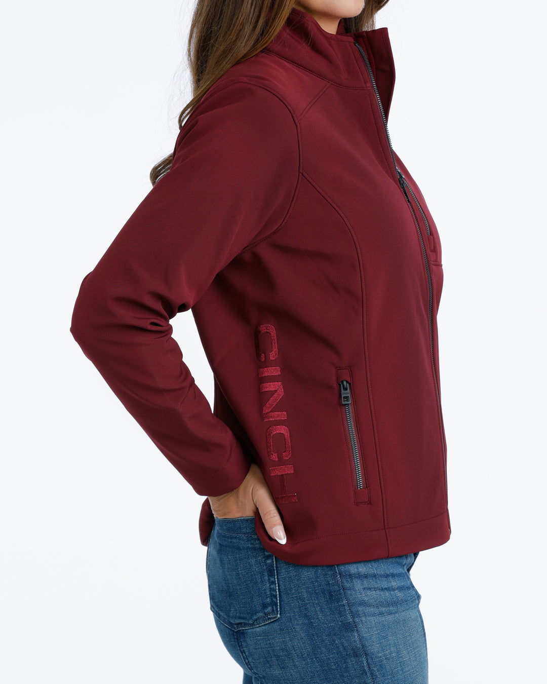 woman wearing a burgundy Cinch jacket with the logo on the hip and blue jeans on a white background