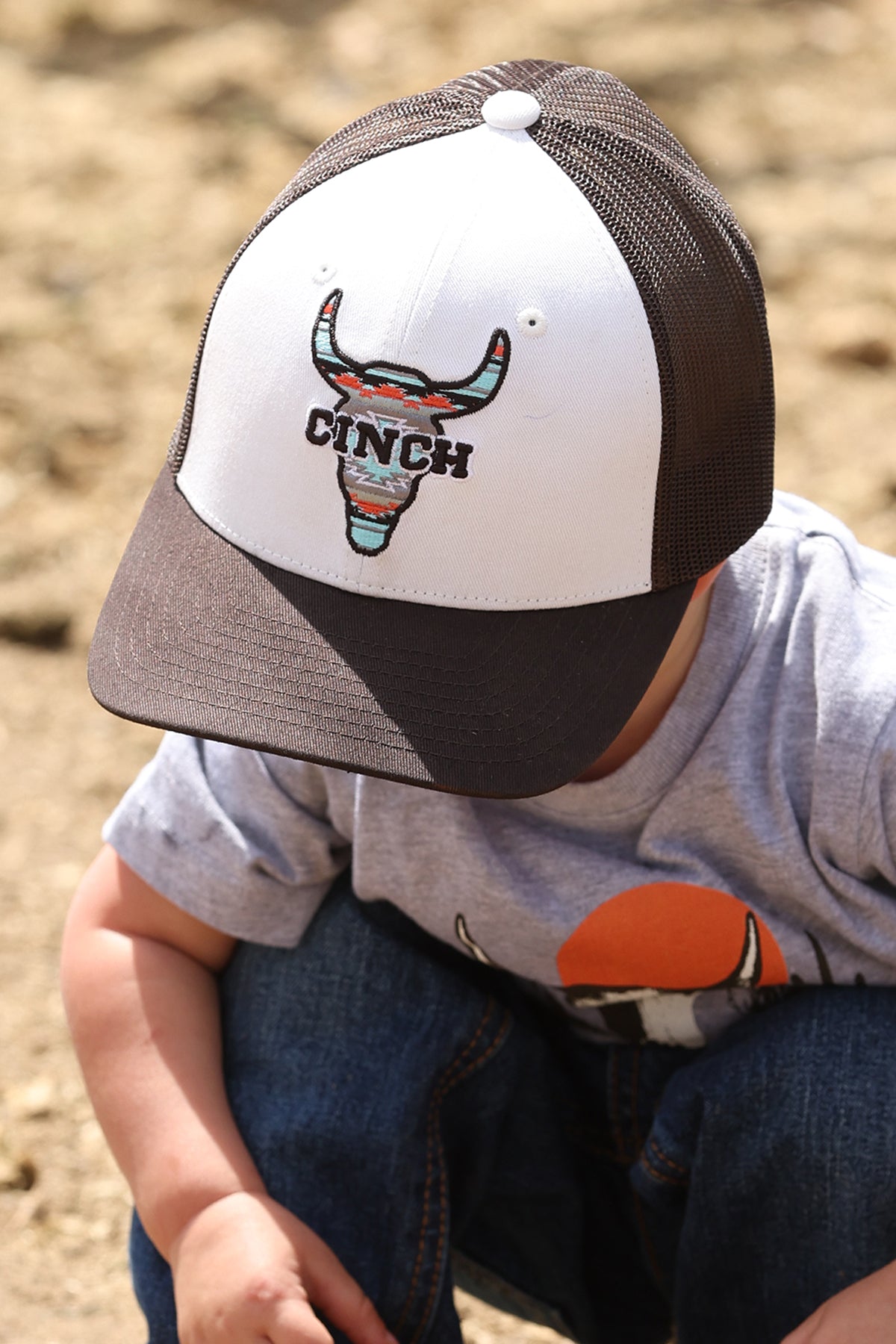 Child wearing a cap with a bull logo and 'Cinch' branding on a dirt background