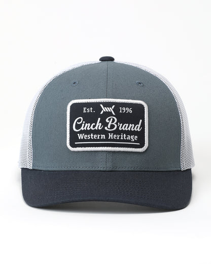 A gray and navy blue trucker hat with white mesh back panels and a black rectangular patch on the front that reads 'Cinch Brand Western Heritage'.
