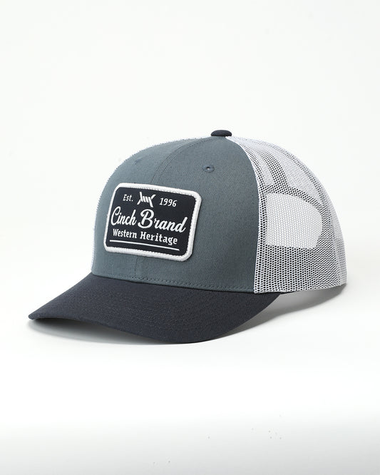 A trucker-style baseball cap with a slate gray front panel, black brim, and white mesh back panels. The front panel features a rectangular patch that reads 'Cinch Brand Western Heritage' with 'Est.