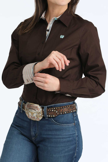 Woman wearing a brown long-sleeve 'Cinch' shirt with a visible brand logo, blue jeans, and a decorative belt on a white background.
